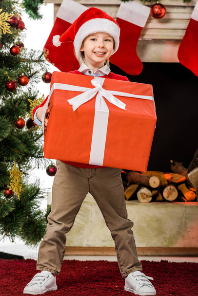 smiling little kid holding big red christmas gift and looking at camera - Foto, Bild