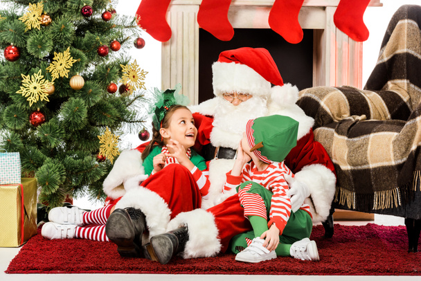 santa embracing happy kids while they sitting on floor near fireplace together - Foto, Imagen