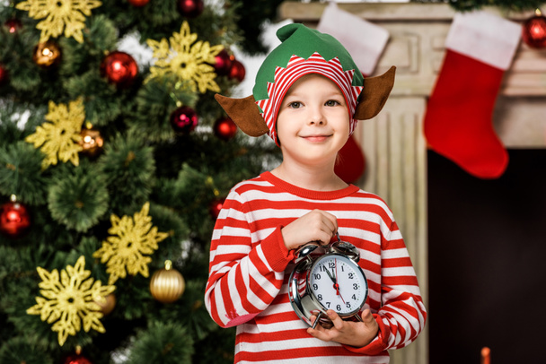 adorable little kid in elf costume with alarm clock standing in front of christmas tree - Foto, Bild