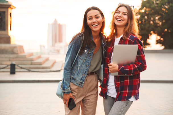 Two attractive young girls friends standing together at the city street, holding laptop and mobile phone, talking - 写真・画像