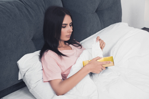 mujer joven enferma tomando servilleta de papel fuera de la caja mientras está sentado en la cama
 - Foto, Imagen