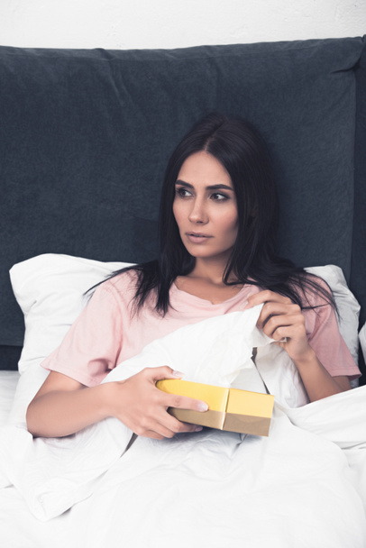 beteg fiatal nő vesz papír szalvéta dobozból ülve, ágyban - Fotó, kép