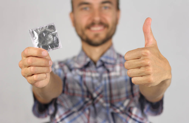 man in plaid shirt holds a new condom in hand, make gesture thumb up, joy and anticipation of pleasure, proper attitude to protection, gray background  - Fotoğraf, Görsel