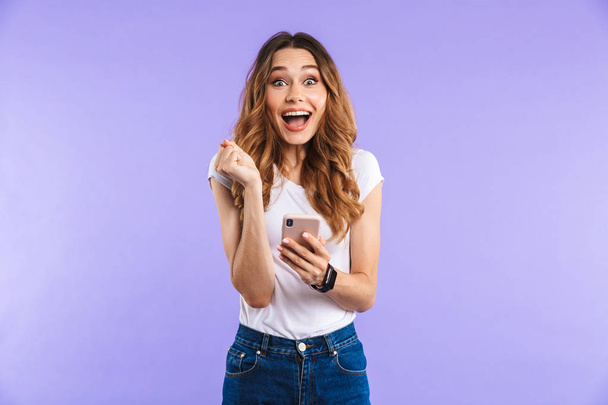 Porträt eines aufgeregten jungen Mädchens, das isoliert vor violettem Hintergrund steht und sein Handy in der Hand hält - Foto, Bild