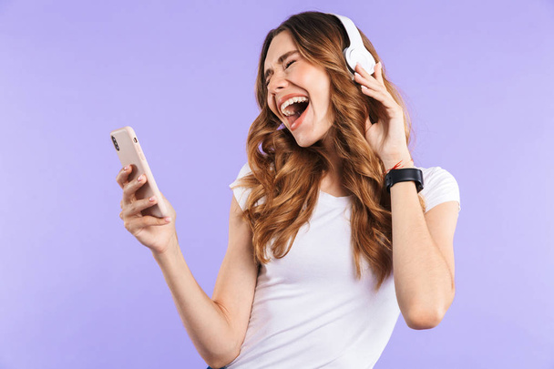 Portrait of a joyful young girl holding mobile phone isolated over violet background, listening to music with headphones - Foto, Imagem