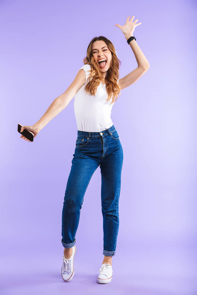 Full length portrait of an energetic young girl holding mobile phone isolated over violet background, listening to music with headphones - Photo, Image