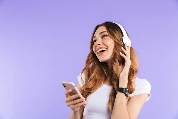 Portrait of a happy young girl holding mobile phone isolated over violet background, listening to music with headphones - Foto, Imagen