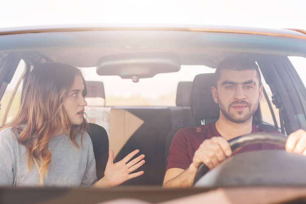 Terrified female gestures actively and tells something emotionally to her husband who drives car and tries not distract from road, have long trip in new automobile. People, family, trip concept - Фото, зображення