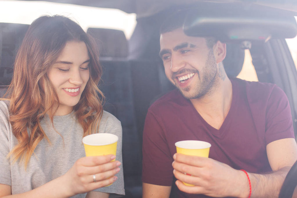 Glad female and male couple have coffee break in car, have long trip, enjoys beautiful views, spend vacation together, reach destination wih high speed. Family has trip or journey by vehicle - Fotó, kép