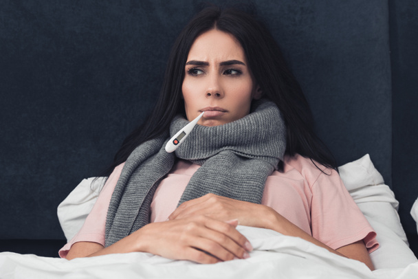 sick young woman measuring temperature with mouth thermometer while lying in bed - Photo, Image