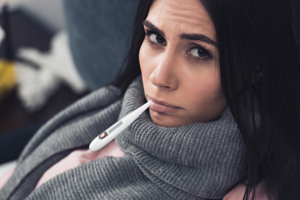 Kranke junge Frau misst Temperatur mit Mundthermometer und blickt in Kamera - Foto, Bild
