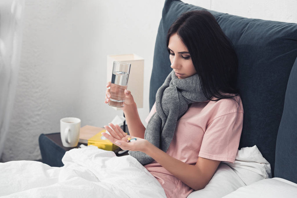 хвора молода жінка тримає таблетки і склянку води, лежачи в ліжку
 - Фото, зображення