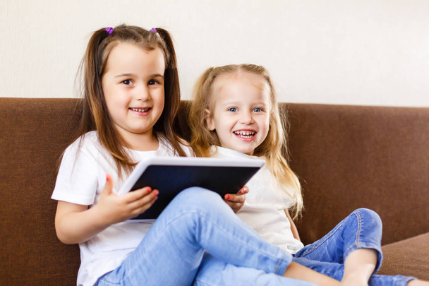 Kaksi suloista pikkusiskoa leikkii digitaalisen tabletin kanssa kotona sohvalla
 - Valokuva, kuva