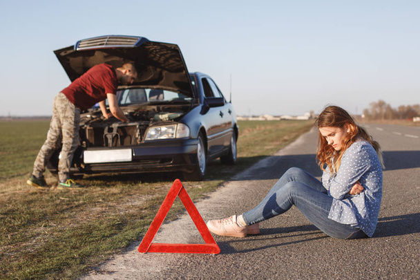 Busy young male driver tries to solve problem with damage engine of car, stands in front of opened hood while his girlfriend sit on road with depressed look, being tired of repairing, need help - Foto, Imagen