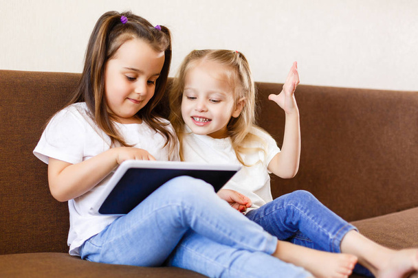 Twee kleine meisjes met behulp van de touchpad en smartphone en zittend op de Bank thuis - Foto, afbeelding