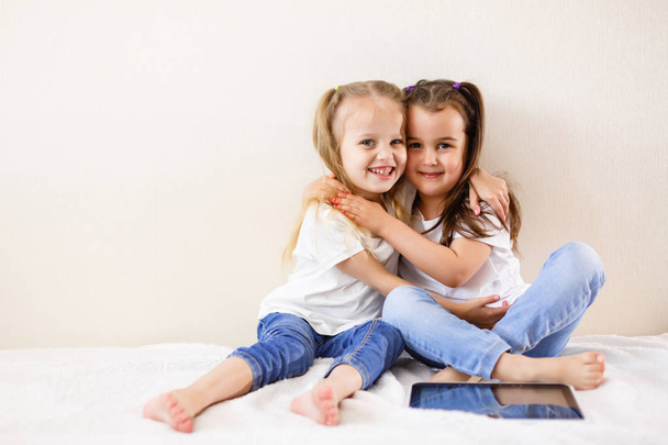 Two happy little girls hugging sitting on bed with digital tablet - Foto, afbeelding