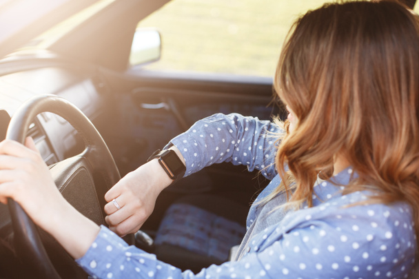 Photo of attractive female checks time on watch, sits in car, stuck in traffic jam and being late for meeting with colleagues, has to delay it. Lovely woman rides auto . People, driving and hurry - Fotoğraf, Görsel