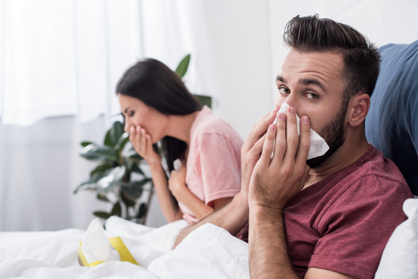 diseased young sick couple sneezing into paper napkins while sitting in bed - Photo, Image