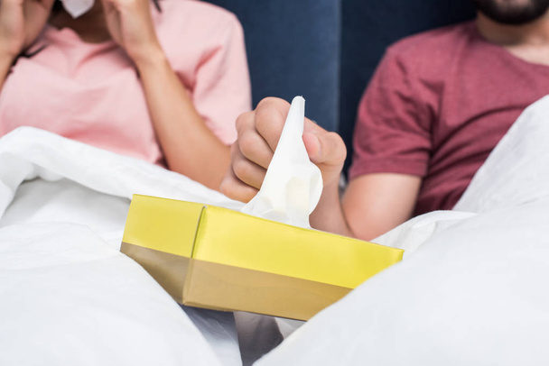 обрезанный снимок больной пары, сидящей в постели и вынимающей бумажные салфетки из коробки
 - Фото, изображение