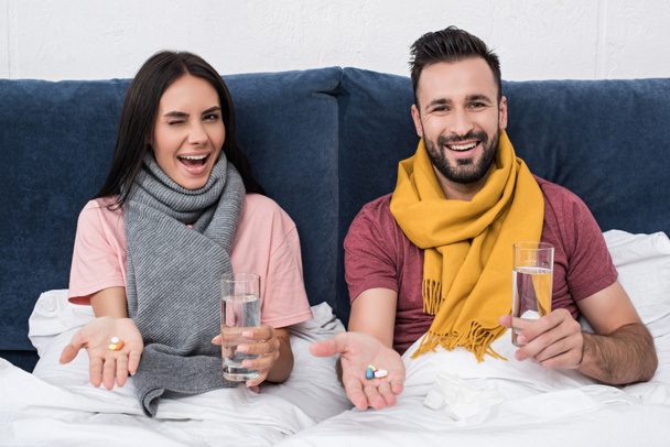 ευτυχισμένο ζευγάρι άρρωστος με χάπια και ποτήρια νερό κάθεται στο κρεβάτι και κοιτάζοντας κάμερα - Φωτογραφία, εικόνα