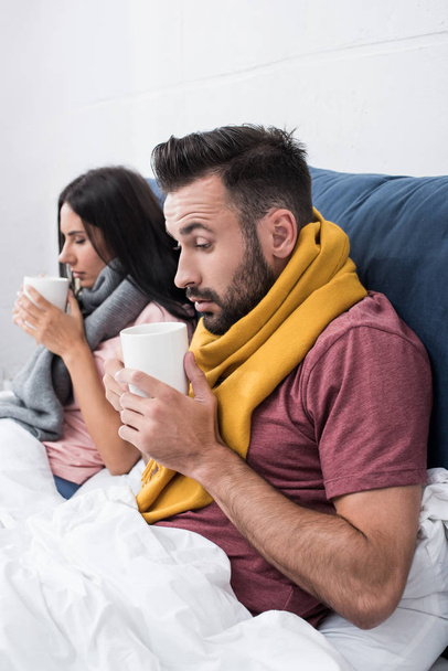 pareja joven enferma en bufandas sentados en la cama con tazas de bebida caliente
 - Foto, Imagen