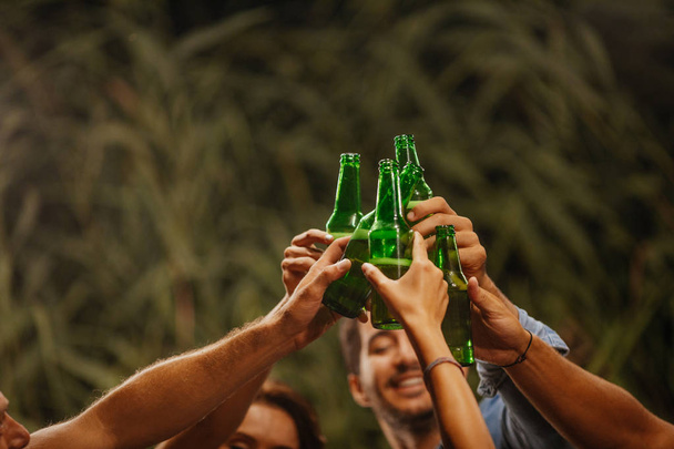 Ręce grupy ludzi nieczytelna opiekania z butelek piwa na zewnątrz impreza. - Zdjęcie, obraz