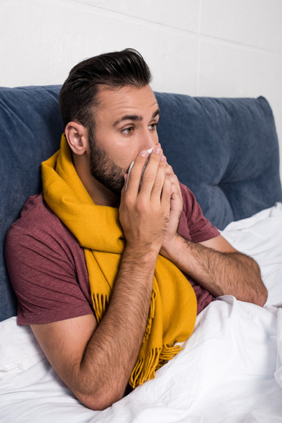 sick young man sneezing while lying in bed - Photo, Image