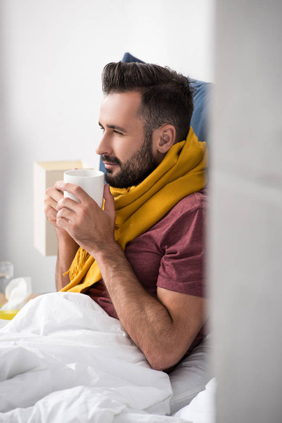 hasta genç adam yatakta otururken sıcak çay tutan eşarp ile gülümseyen - Fotoğraf, Görsel