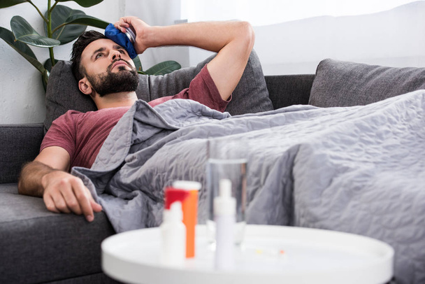 sick young man lying on couch and holding ice pack on forehead - Photo, Image