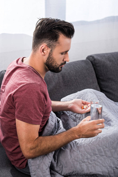 sairas nuori mies istuu sohvalla lasi vettä ja pillereitä
 - Valokuva, kuva