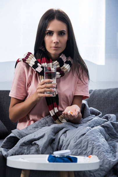 nemocný mladá žena s sklenice vody a prášky sedí na gauči a při pohledu na fotoaparát - Fotografie, Obrázek