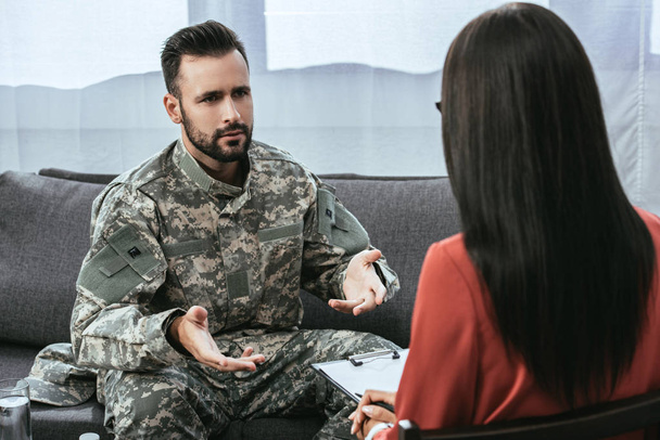 sad soldier with ptsd talking at psychiatrist and gesturing while sitting on couch during therapy session - Photo, Image