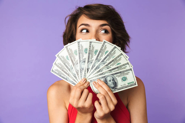 Happy Pretty brunette woman covering her face with money and looking away over purple background - Fotó, kép