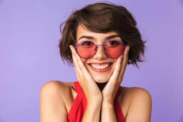 Happy Pretty brunette woman in sunglasses touching her cheeks and looking at the camera over purple background - Fotoğraf, Görsel