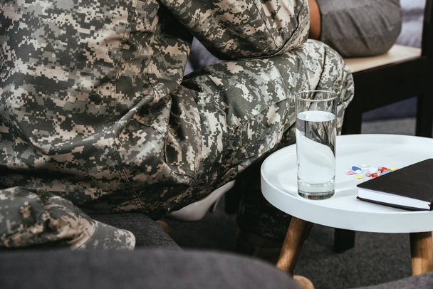 cropped shot of soldier sitting on couch with glass of water and pills on table - Foto, imagen