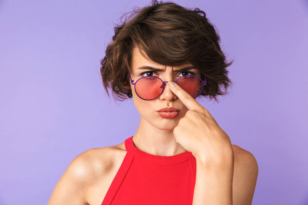 Serious Pretty brunette woman corrects her sunglasses and looking at the camera over purple background - Foto, afbeelding