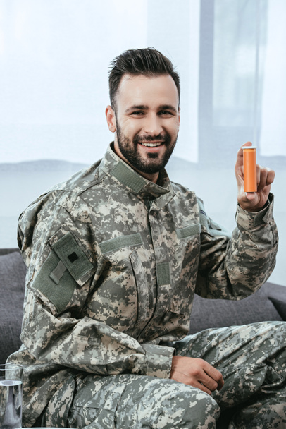 счастливый молодой солдат держит пластиковую банку с таблетками и смотрит в камеру
 - Фото, изображение