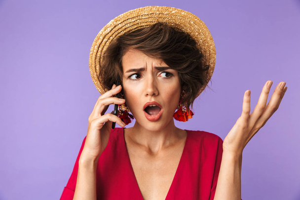Confused Pretty brunette woman in dress and straw hat talking by smartphone while looking away over purple background - Foto, afbeelding