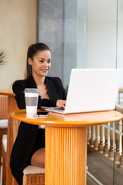 Smiling elegant businesswoman with coffee sitting at high table and surfing laptop - Photo, Image
