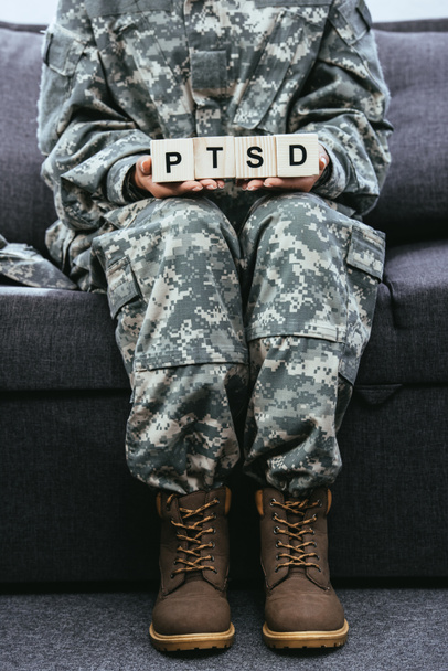 cropped shot of female soldier in military uniform sitting on couch and holding wooden cubes with PTSD sign - Photo, Image