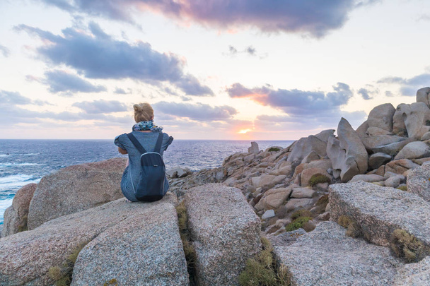 一人の若い女性の旅行者の時計カポテスター、サルデーニャ、イタリアの壮大な岩の美しい夕日. - 写真・画像