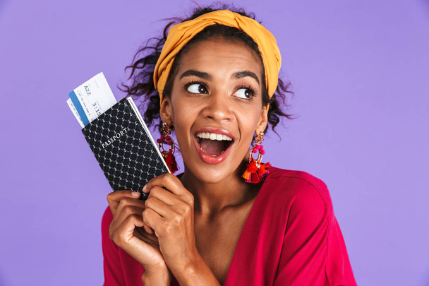 Playful african woman in dress holding passport with tickets while looking away over purple background - Φωτογραφία, εικόνα