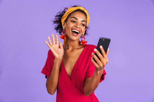 紫背景にカメラを振りながらスマート フォンでビデオ通話で話しているドレスでうれしそうなアフリカ女性 - 写真・画像