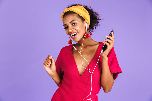 Happy african woman in dress and earphones listening music by smartphone while looking at the camera over purple background - Fotografie, Obrázek