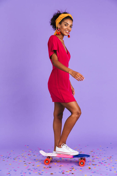 Full length portrait of a smiling young african woman in headband standing on skateboard over violet background - Fotografie, Obrázek