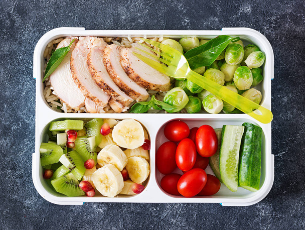 Здоровый контейнер для приготовления зеленой еды с куриным филе, рисом, брюссельской капустой, овощами и фруктами на сером камне
 - Фото, изображение