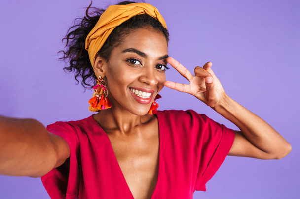 Cheerful african woman in dress making selfie while showing peace gesture and looking at the camera over purple background - Фото, зображення