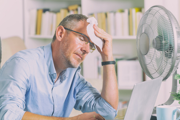 Человек страдает от жары во время работы в офисе и пытается остыть за счет вентилятора - Фото, изображение