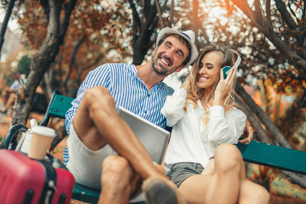 Pareja escuchando música con una tableta sentada en un banco en un parque urbano
 - Foto, Imagen