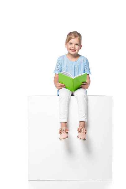 child with book sitting on white cube isolated on white - Zdjęcie, obraz